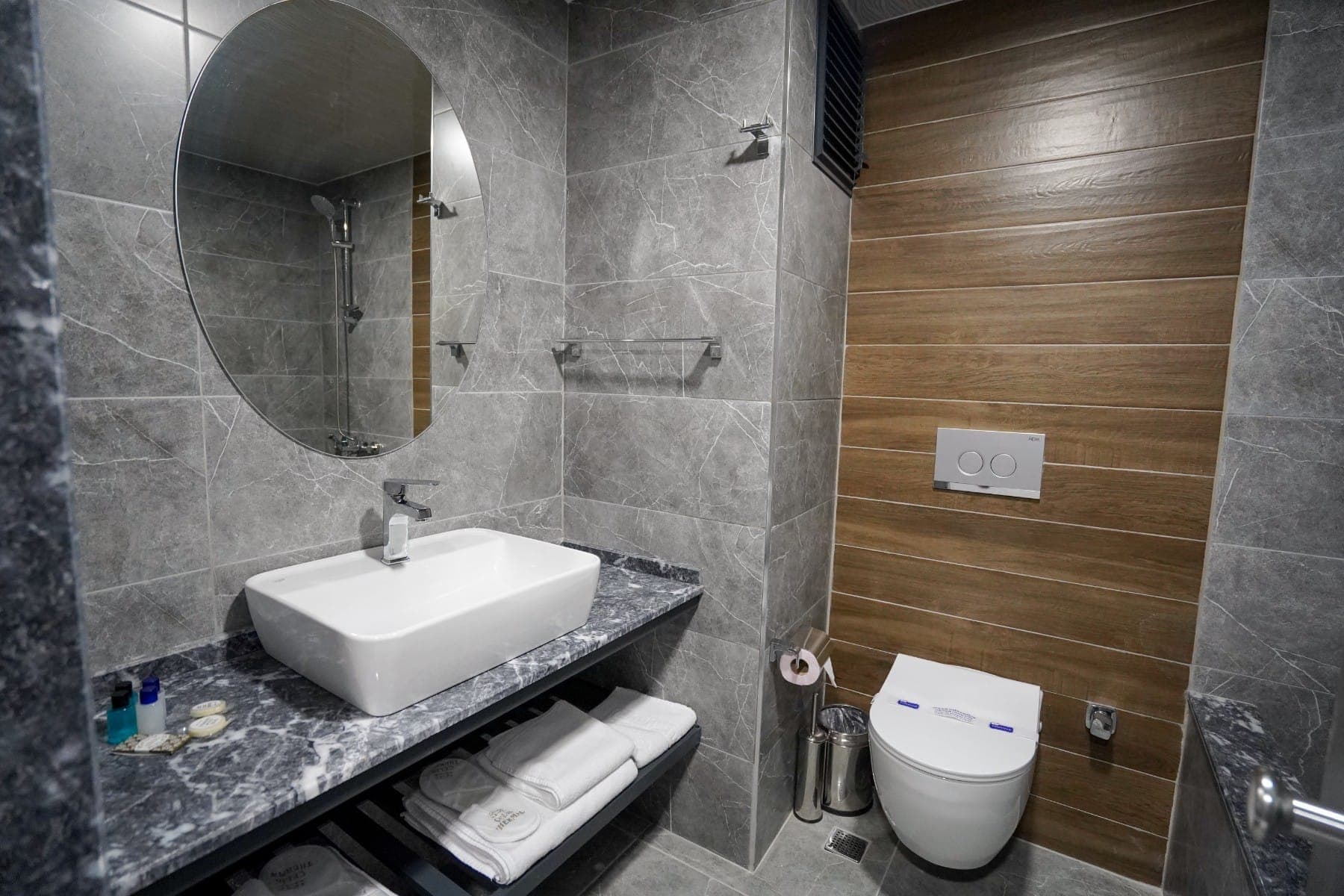 Gri mermer banyoda oval ayna, tezgâh üstü lavabo ve katlanmış havlular
